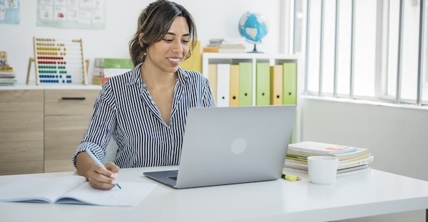 Teacher at classroom sitting at desk and using laptop. She is talking with students on video call.